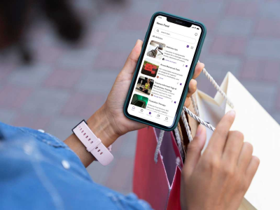 Image of a woman’s holding a phone with the home screen of Fem Entity’s app. The phone’s screen is visible over her shoulder while she is holding shopping bags.